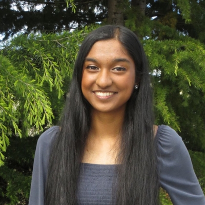 Nayha Auradkar, a South Asian woman with brown skin and long black straight hair, smiling at the camera. She is wearing a gray blouse and there are dark green trees behind her
