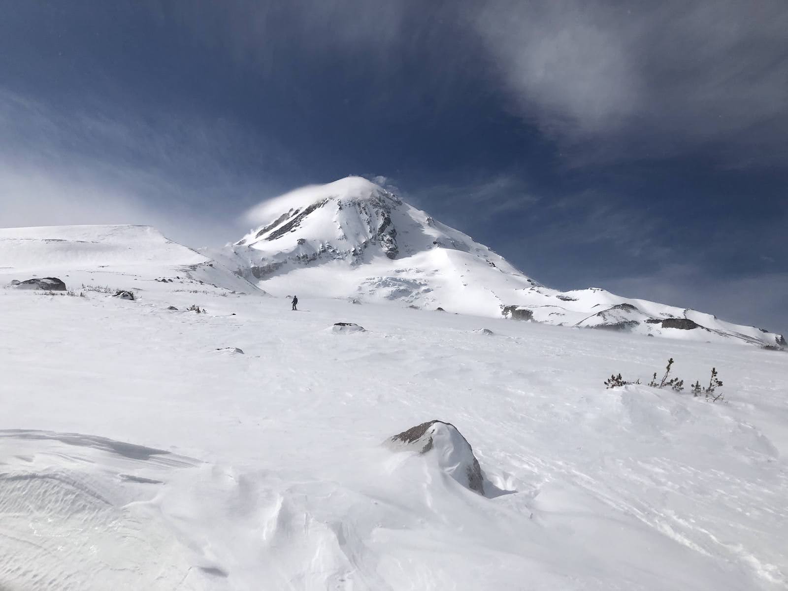 a view of Mt Hood from the Cooper Spur Shelter; a skiier descends towards the shelter on a windy snow field