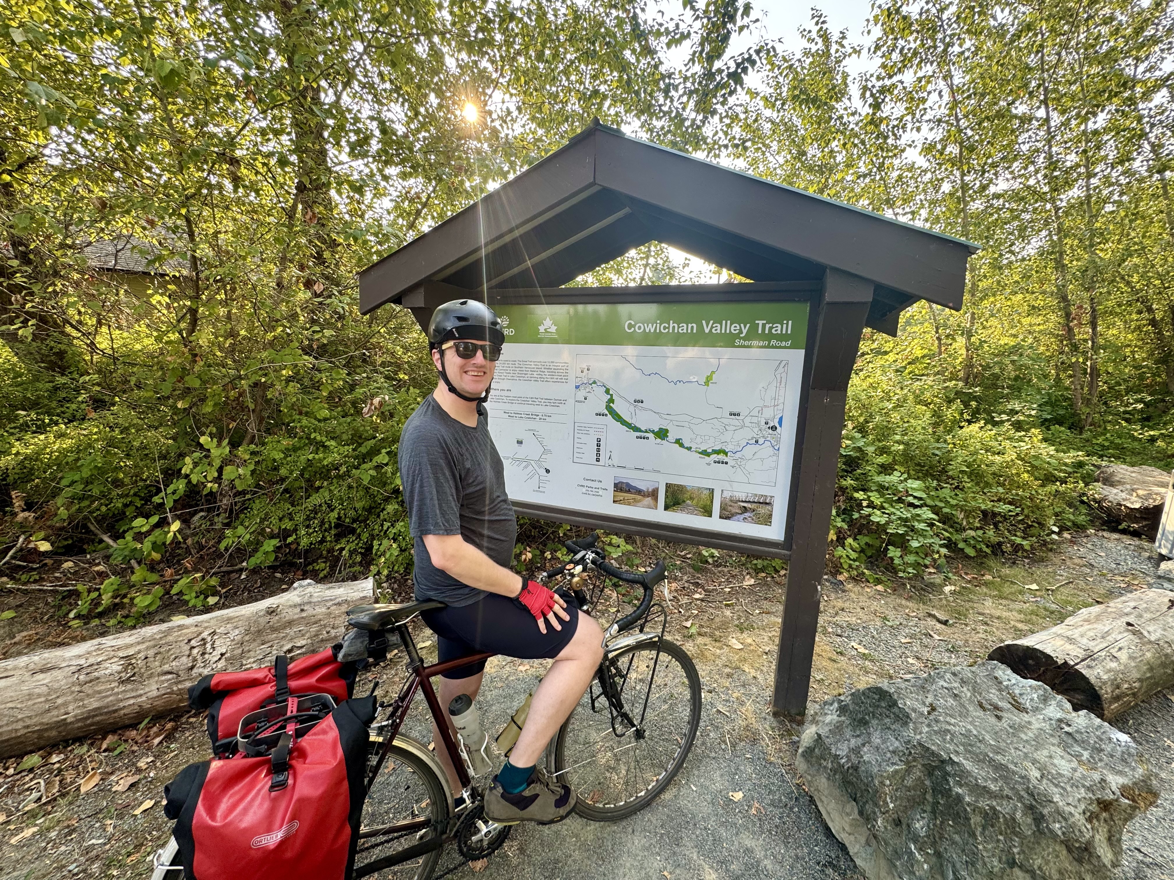man riding bicycle with two panniers stands smiling in front of a sign for the cowichan valley trail